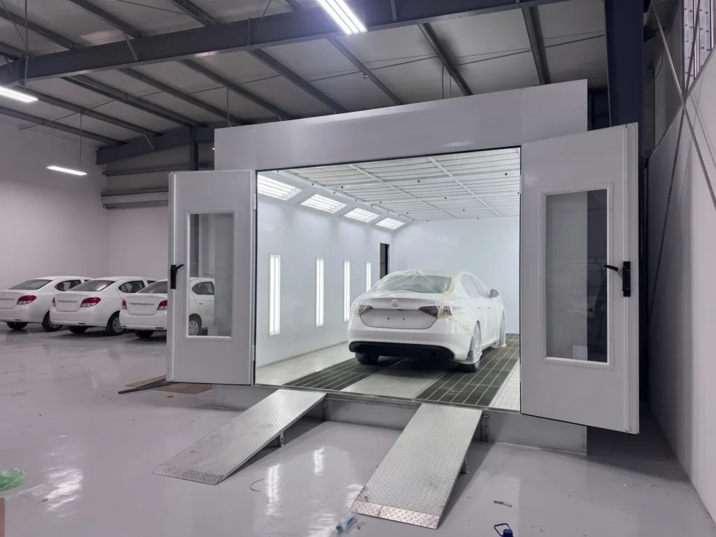 A car wrapped in film for a spot repair inside a paint booth with open doors at a rental company