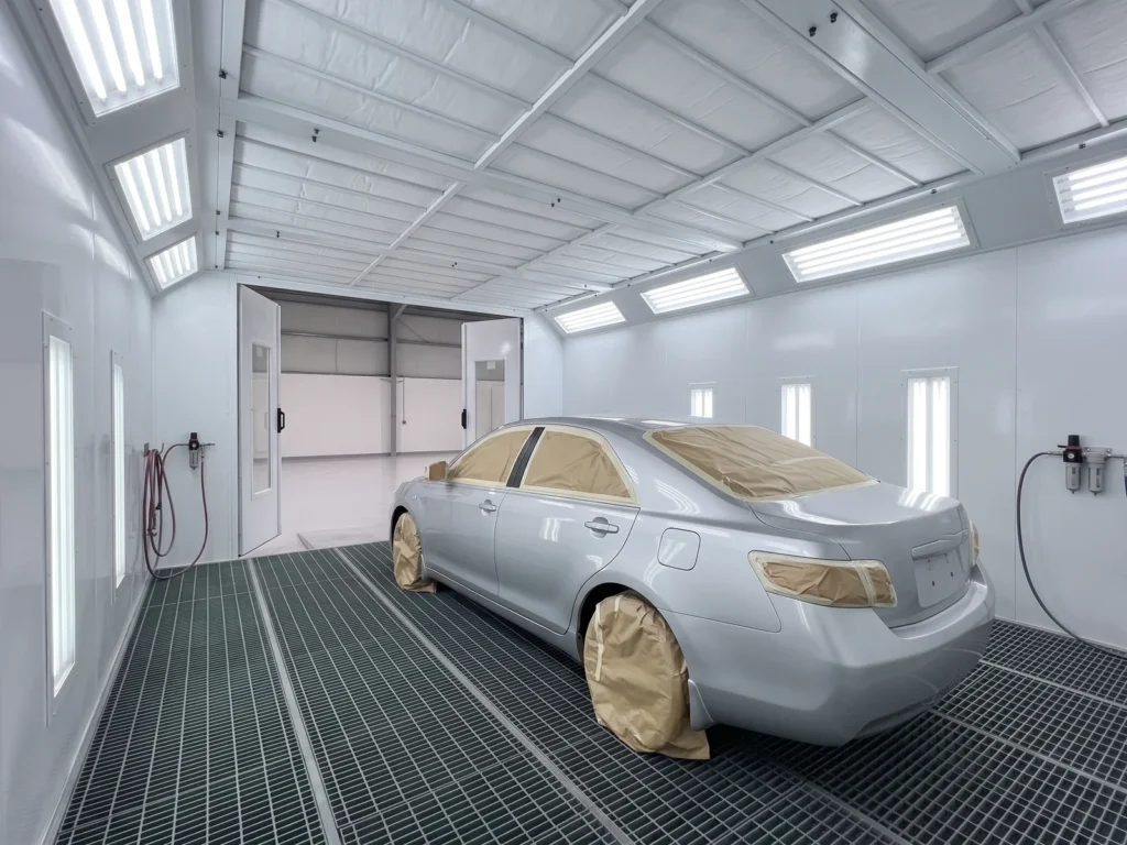 Silver car with masked windows and wheels inside a paint booth at a collision repair centre