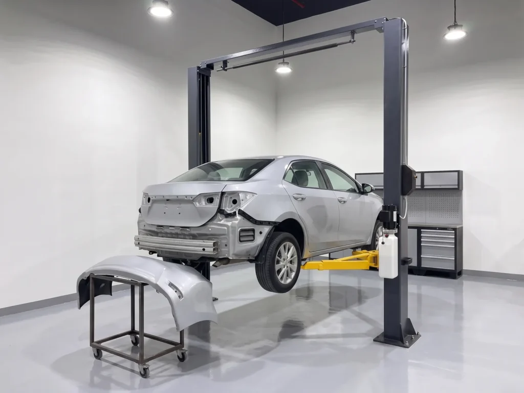Silver car with its rear bumper removed on a two-post lift, the bumper on a trolley beside it