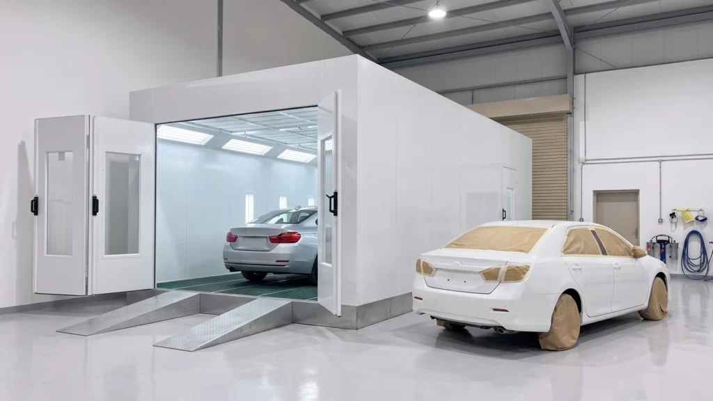 A painted car inside a paint booth with open doors and a second masked car waiting its turn