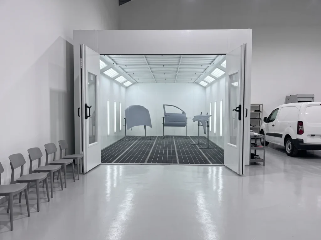 Car body panels in primer on stands inside a paint booth with open doors in a training workshop